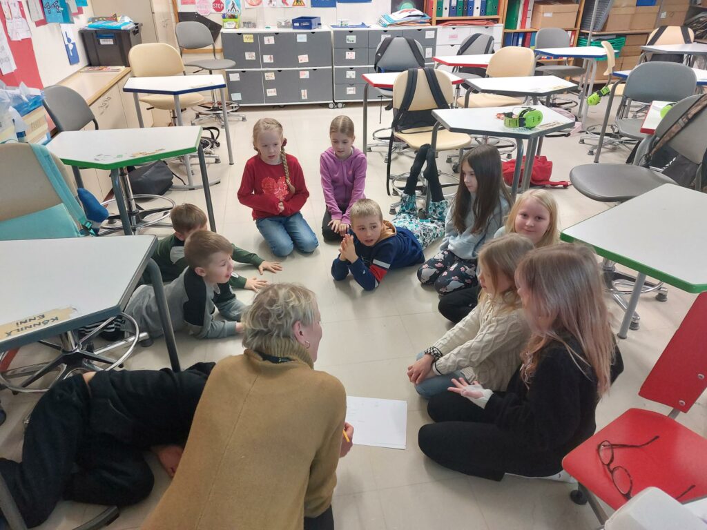 Lapsia sekä opettaja istumassa luokkahuoneen lattialla pienessä piirissä.