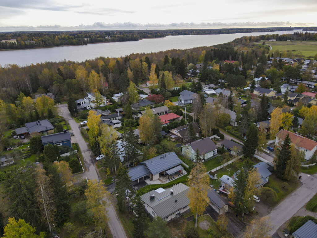 Ilmakuva syksyisestä järvimaisemasta, jossa etualalla näkyy pientaloalue, vehreitä pihoja ja ruskaväreihin pukeutuneita puita. Talot ovat omakotitaloja, ja alueella on hiljainen, luonnonläheinen tunnelma. Taustalla näkyy järvi, jonka rannat ovat metsän peitossa, ja taivaalla leijuu harmaita pilviä, jotka korostavat viileää syksyn ilmaa.