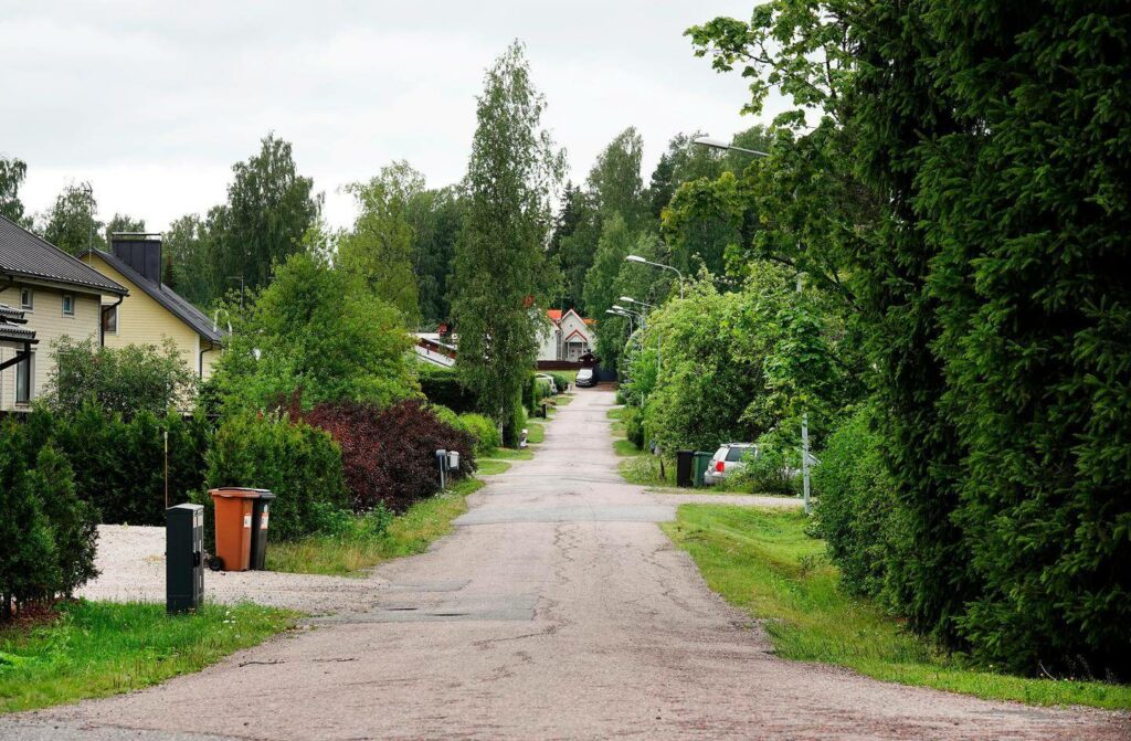 Kuvassa on vehreä ja rauhallinen asuinalue, jossa on kapeahko asfalttitie, jota reunustavat omakotitalot ja runsaasti kasvillisuutta. Talot ovat osittain piilossa pensaiden ja puiden takana. Kadun varrella näkyy roskalaatikoita ja muutamia pysäköityjä autoja. Tien varrella on katulamppuja, ja taustalla häämöttää yksi suurempi valkoinen talo punaisella katolla. Ympärillä on paljon vihreää, ja taivas näyttää olevan pilvinen.