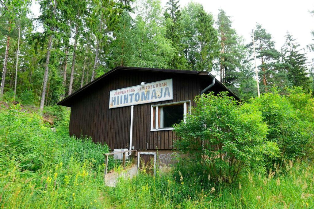 Kuvassa on ruskea puurakennus, jonka julkisivussa lukee "Järvenpään Hiihtoseuran Hiihtomaja" vanhassa kyltissä. Rakennus sijaitsee metsän keskellä, ja sen ympärillä kasvaa runsaasti korkeaa ruohoa ja pensaita. Taustalla näkyy korkeita havupuita, ja sää näyttää pilviseltä mutta kirkkaalta. Rakennuksen ikkunat ja ulko-osa vaikuttavat hieman kuluneilta.